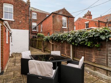 A patio area with a black glass table and four chairs with white cushions surrounded by brick buildings and a wooden fence with green ivy at Elkhorn in Southwold