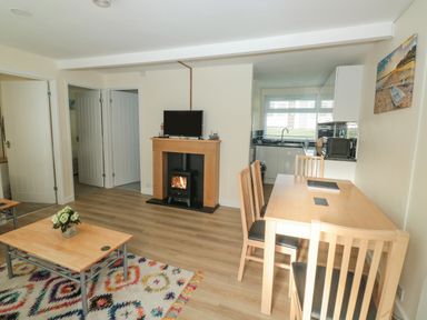 A room with wooden dining table and chairs a coffee table with flowers a fireplace with a stove and a television and a kitchen area at The Bolthole in Dartmouth