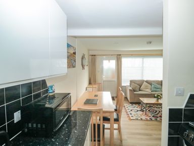 A kitchen with a microwave and black tiled backsplash next to a dining table with four chairs leading to a living room with a beige sofa and coffee table at The Bolthole in Dartmouth
