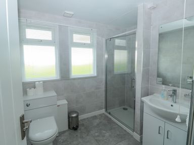 A bathroom with a toilet, shower with glass door, sink with mirror, and two windows with blinds at The Bolthole in Dartmouth