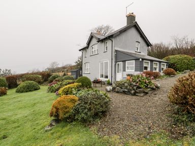 A two-story house with a grey exterior surrounded by a garden with shrubs and a gravel pathway at Sibrwd Y Mor in Harlech