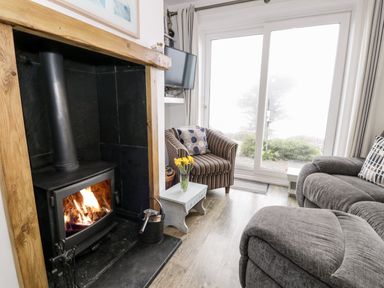 A living room with a wood burning stove fire a striped armchair a small table with flowers and sliding glass doors at Sibrwd Y Mor in Harlech