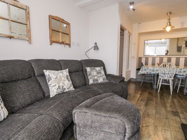 A living room with a gray sofa and ottoman and a dining area with a table with a printed tablecloth at Sibrwd Y Mor in Harlech