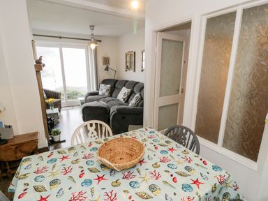 A dining table with a sea-themed tablecloth and a wicker basket with a living room and grey sofa in the background at Sibrwd Y Mor in Harlech