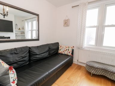 A black leather sofa with patterned cushions next to a window with white curtains and a patterned bench on a wooden floor at Sibrwd Y Mor in Harlech
