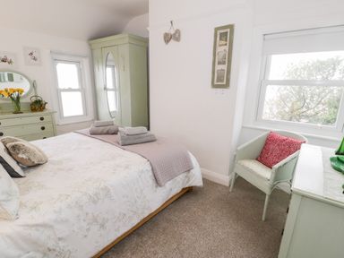 A bedroom with a bed with folded towels bedside table with flowers green wardrobe and chair by window at Sibrwd Y Mor in Harlech