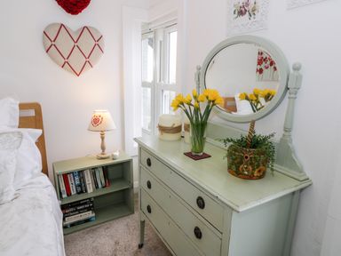 A bedroom corner with a wooden bed frame a small bookshelf lamp and a dresser with a mirror and flowers at Sibrwd Y Mor in Harlech
