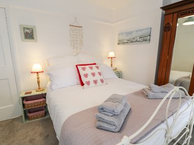 A bedroom with a white bed frame gray towels on a blanket two bedside tables with lamps a wooden wardrobe and wall art at Sibrwd Y Mor in Harlech