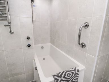 A bathtub with a towel on the edge and tiled walls with a shower handle and grab bar at Sibrwd Y Mor in Harlech