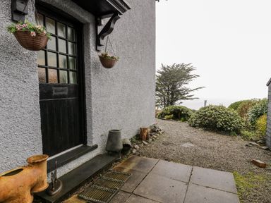 An outdoor entrance with a black door and hanging flower baskets leading to a gravel path and garden at Sibrwd Y Mor in Harlech