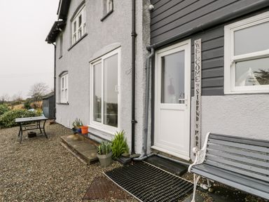 An exterior view of a building with a white door a welcome sign plants on the ground a bench and a table at Sibrwd Y Mor in Harlech
