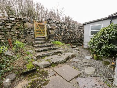 An outdoor stone pathway and steps leading to a wooden gate beside a stone wall and shrubbery at Sibrwd Y Mor in Harlech