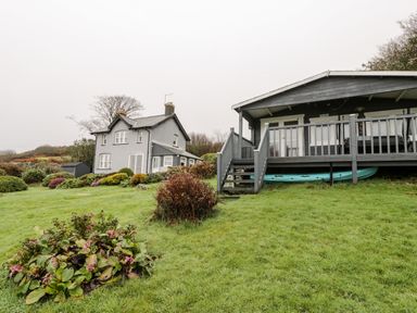 A gray house and a gray cabin with a deck in a grassy garden with shrubs at Sibrwd Y Mor in Harlech