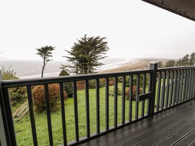 A deck with wooden railing overlooking grassy yard trees and a beach at Sibrwd Y Mor in Harlech
