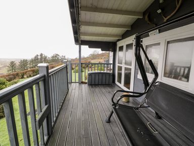 A covered porch with a black swing bench a storage box and a view of trees and a garden at Sibrwd Y Mor in Harlech