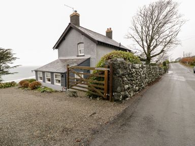 A small house with a stone wall and wooden gate next to a road with leafless trees at Sibrwd Y Mor in Harlech