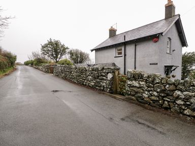 A grey house with two chimneys behind a stone wall and wooden gate beside a wet road at Sibrwd Y Mor in Harlech