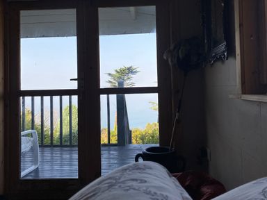 A view through glass doors to a wooden deck with a white chair and greenery beyond inside a bedroom at Sibrwd Y Mor in Harlech