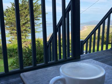 A cup of coffee on a wooden table with a view of a green garden trees water and hills beyond railing at Sibrwd Y Mor in Harlech