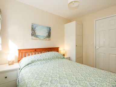 A bedroom with a bed with a patterned green and white bedspread two white bedside tables with lamps a white wardrobe and a painting above the headboard at 11 The Dell in Mundesley
