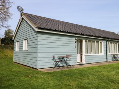 An exterior view of a single-story house with light blue siding and a tiled roof with two small tables and four chairs outside at 11 The Dell in Mundesley