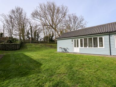 A green grassy yard with leafless trees and a blue single-story building with white doors and windows at 11 The Dell in Mundesley