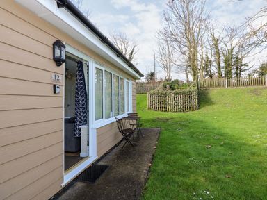An exterior view of a beige house with an open door showing a glimpse of the inside and a small table with two chairs on a concrete patio next to a grass lawn at 12 The Dell in Mundesley