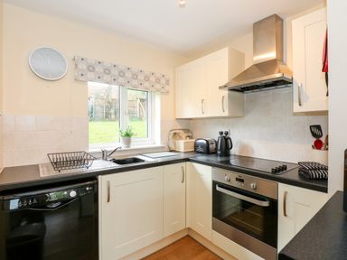 A kitchen with black countertops white cabinets a stove a toaster a kettle a dish rack a clock and a window with a potted plant at 12 The Dell Mundesley