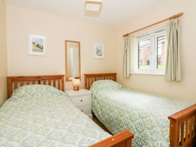 A bedroom with two single beds a white nightstand a lamp a mirror and a window with curtains at 12 The Dell in Mundesley