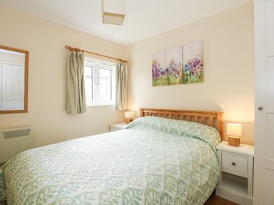 A bedroom with a double bed green patterned bedding bedside tables lamps a window and wall art at 12 The Dell in Mundesley