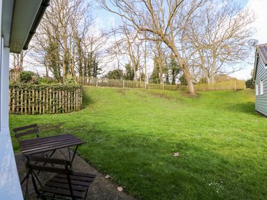A grassy backyard with wooden chairs and table near the house and a wooden fence with trees at 12 The Dell in Mundesley