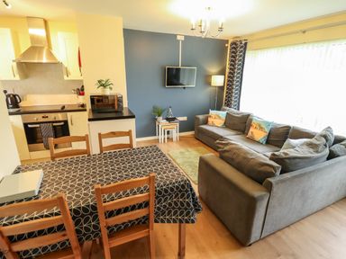 A living room with grey sofas a small green rug and a wall-mounted tv next to a dining table with four wooden chairs and a kitchen area at 12 The Dell in Mundesley