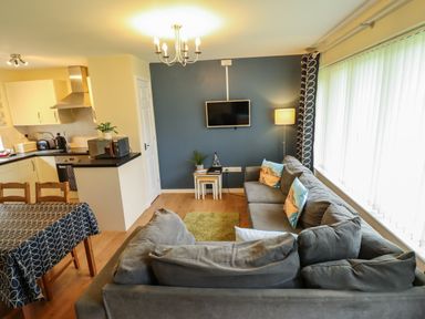 A living room with a gray sofa a wall-mounted TV small tables and a kitchen area at 12 The Dell in Mundesley
