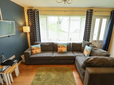 A living room with a grey sectional sofa and decorative cushions by a window with vertical blinds and patterned curtains at 12 The Dell in Mundesley