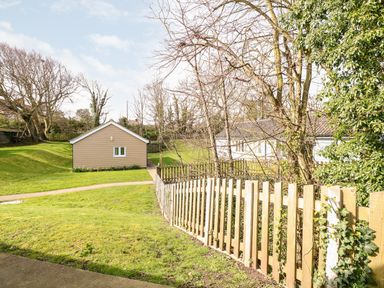 A garden area with a wooden fence a small building and a larger white house surrounded by trees at 15 The Dell in Mundesley