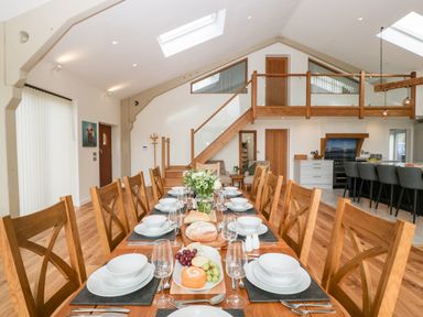A dining area with a long wooden table set with plates, bowls, glasses, cutlery, bread, and fruit with a staircase and kitchen in the background at Bilsham Meadows in Aust