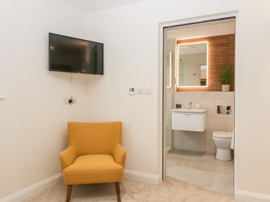 A yellow armchair below a wall-mounted television next to an open door leading to a bathroom with a lit mirror a sink and a toilet at Bilsham Meadows in Aust