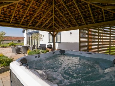 An outdoor hot tub under a wooden gazebo with patio furniture and a barbecue outside a house at Bilsham Meadows in Aust