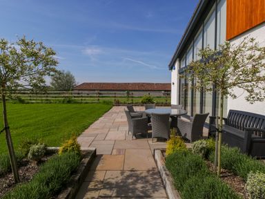 An outdoor patio area with a round table and six chairs next to a lawn and a building with large windows at Bilsham Meadows in Aust