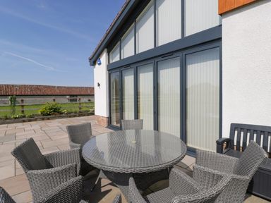 An outdoor patio area with a round glass table surrounded by six wicker chairs near a building with large windows at Bilsham Meadows in Aust
