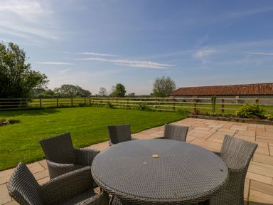 An outdoor patio with a round table and six chairs next to a grassy lawn and wooden fence at Bilsham Meadows in Aust