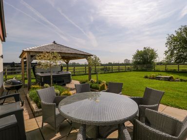 An outdoor patio with a round glass table and wicker chairs next to a hot tub under a wooden gazebo at Bilsham Meadows in Aust