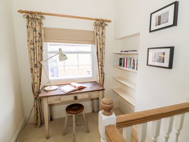 A small study area with a wooden desk and stool by a window with curtains and built in shelves with books at 4 The Old Council House Shipston-On-Stour
