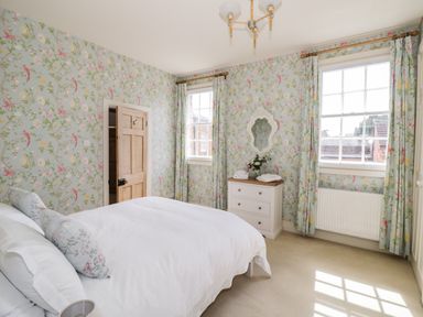 A bedroom with a bed white bedding floral wallpaper two windows with floral curtains a chest of drawers with a mirror and flowers at 4 The Old Council House Shipston-On-Stour