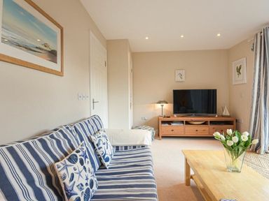A living room with a striped sofa and patterned cushions a wooden TV cabinet a coffee table with flowers and framed pictures on the walls at Smugglers Cove 7 Beadnell