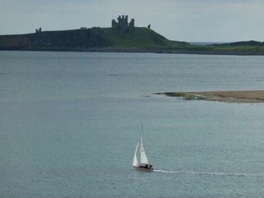 A sailboat on the water with a grassy hill and castle ruins in the background at Smugglers Cove 7 in Beadnell
