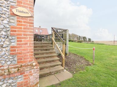 Exterior stone and brick wall with a sign that says Timbers stairs with wooden railing leading to a terrace with chairs and a table green field and trees in the background at Timbers Weybourne