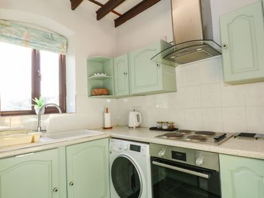 A kitchen with light green cabinets a washing machine an oven a stovetop and a window with a floral blind at Timbers in Weybourne