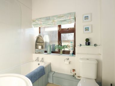 A bathroom with a bathtub a toilet a window with a patterned blind and wooden boat-shaped shelf at Timbers in Weybourne