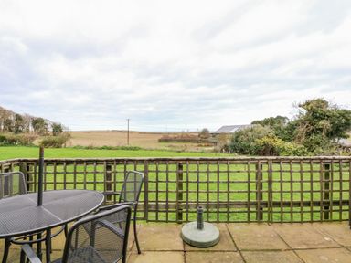 A patio with a metal table and chairs overlooking a grassy field and distant buildings at Timbers in Weybourne
